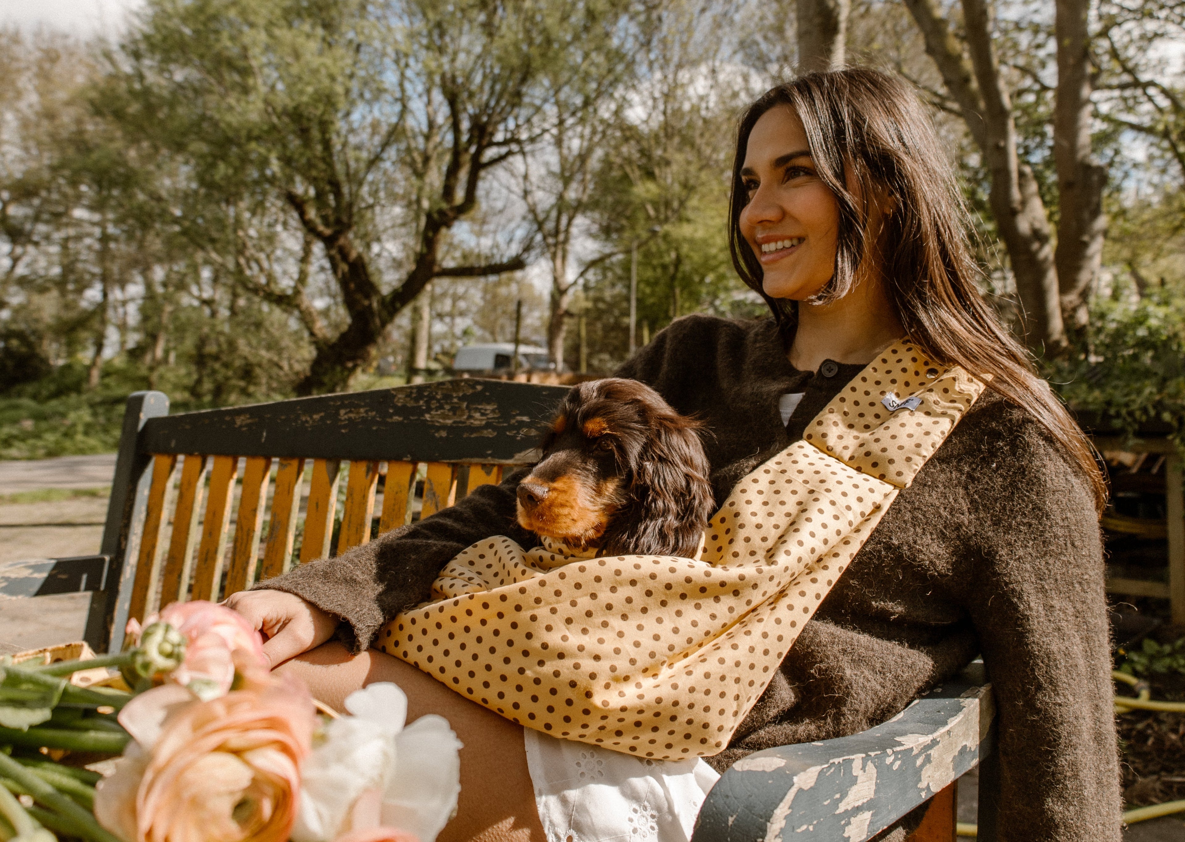 Woman sitting on a bench with a dog, holding flowers, in a park setting. Polka dot leash, polka dot riem en halsband en draagzak voor honden